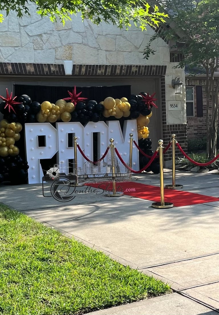 Large white PROM letters decorated with black and gold balloons on a house garage, with red carpet and velvet rope barriers in front