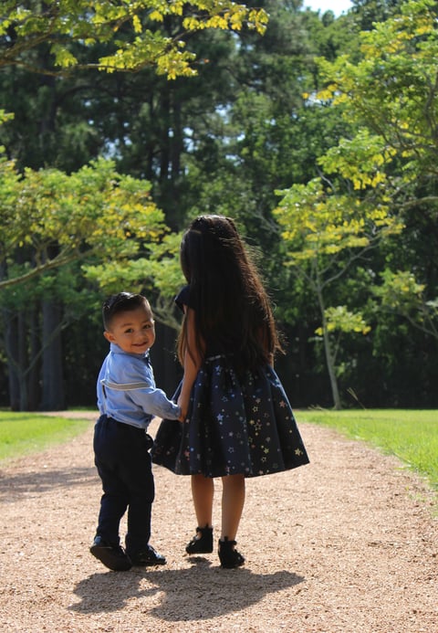Young girl in dark dress holding hands with toddler boy on tree-lined dirt path in park