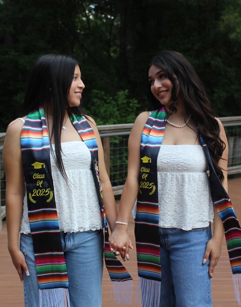 Two young women wearing Class of 2025 graduation stoles with colorful striped patterns, white lace tops, and jeans holding hands outdoors