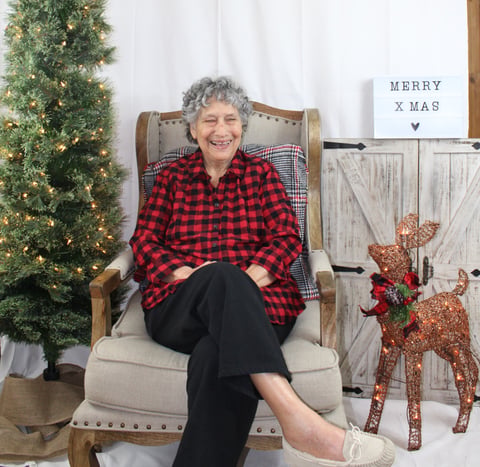 Elderly woman with gray hair smiling in chair wearing red plaid shirt, surrounded by Christmas decorations including garland and reindeer