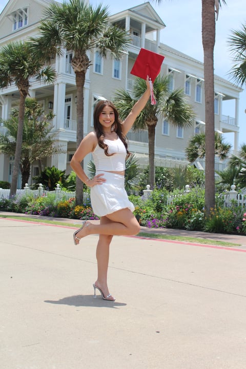 Young woman in white outfit holding red graduation cap celebrating in front of coastal mansion with palm trees