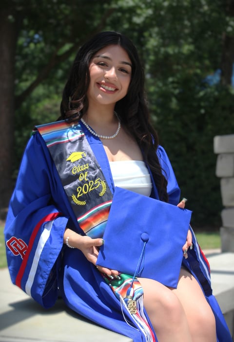 Young woman in blue graduation gown with Class of 2025 sash holding diploma, smiling outdoors with green foliage background