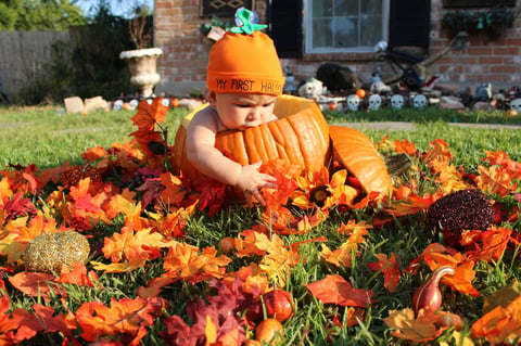 Baby in pumpkin costume sitting among fall flowers and pumpkins in front yard