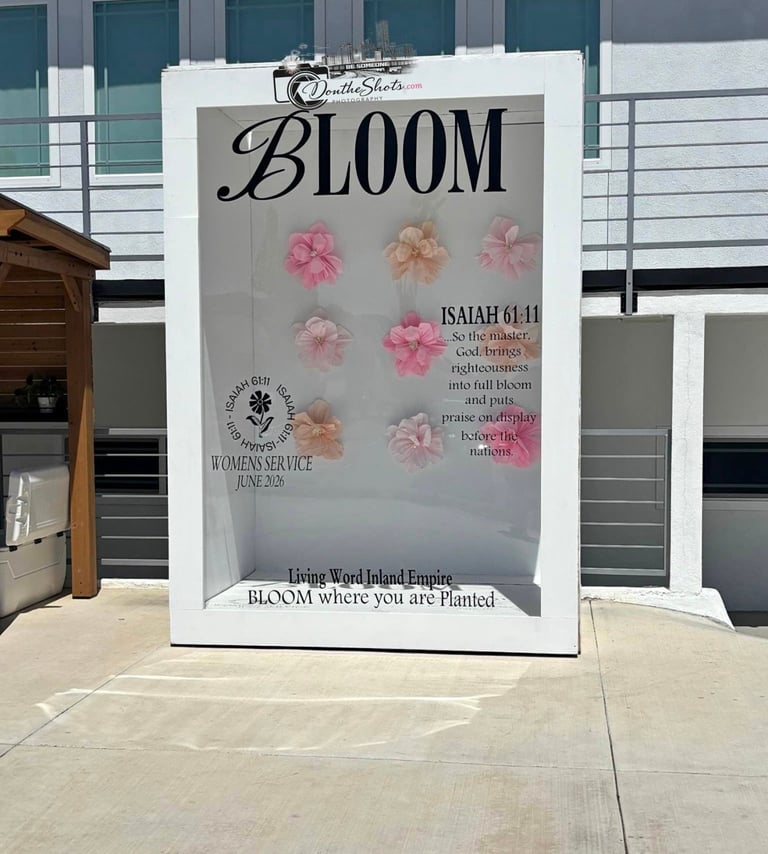 White outdoor sign displaying BLOOM with pink and coral flowers and Isaiah 61:11 scripture verse at a womens service event
