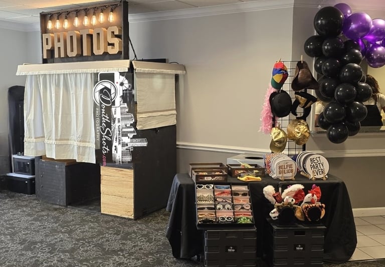 Photo booth setup at event with illuminated PHOTOS sign, white curtain, and nearby display table with balloons and props