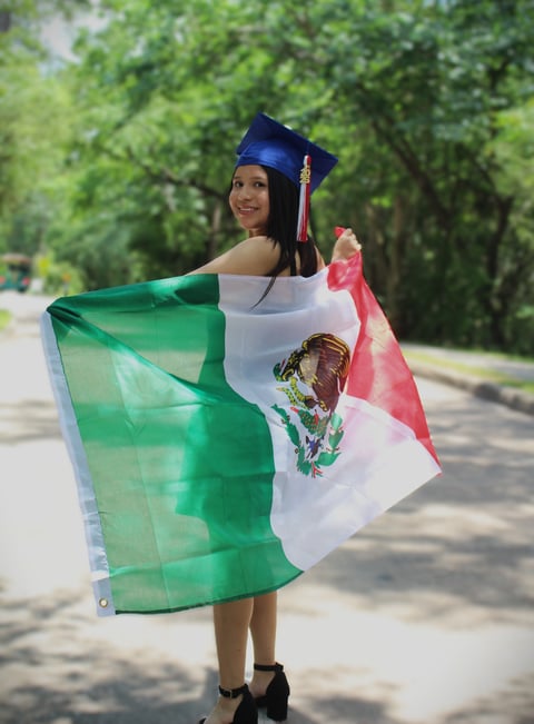 Young graduate wearing blue cap and gown holding Mexican flag outdoors on tree-lined street