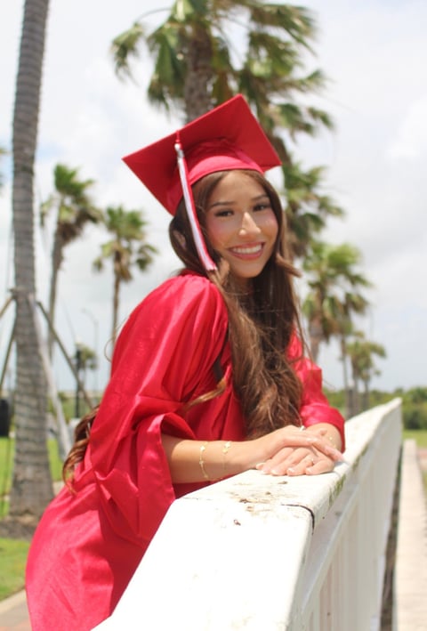 Graduate in red cap and gown smiling while leaning on a railing with palm trees in the background