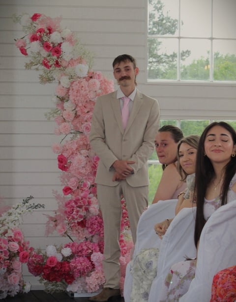 Man in beige suit beside pink and white floral wall with bridesmaids
