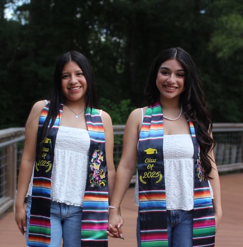 Two teenage girls wearing matching white tank tops and colorful striped graduation stoles with Class of 2025 embroidery, standing together outdoors holding hands