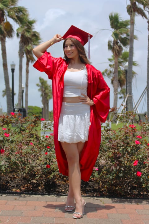 Woman in red graduation gown and cap posing in front of palm trees and red flowers