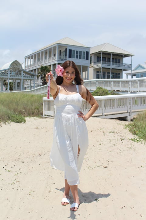 Woman in white dress holding pink flowers on beach with coastal houses in background