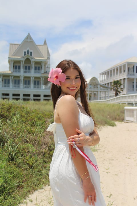 Woman in white dress with pink flower in hair standing on beach near Victorian buildings