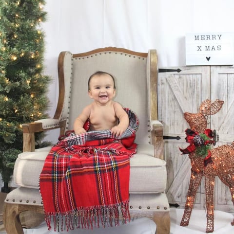 Smiling baby sitting in wingback chair wrapped in red plaid blanket with Christmas decorations and Merry Xmas sign in background
