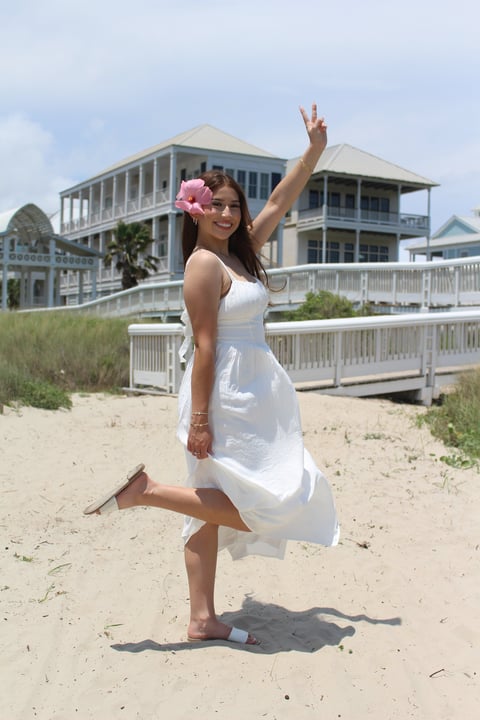 Woman in white dress with pink flower in hair posing on beach in front of large beach house