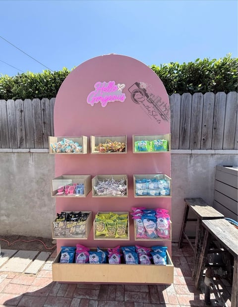 Pink vending stand with shelves displaying snacks and beverages in a backyard setting