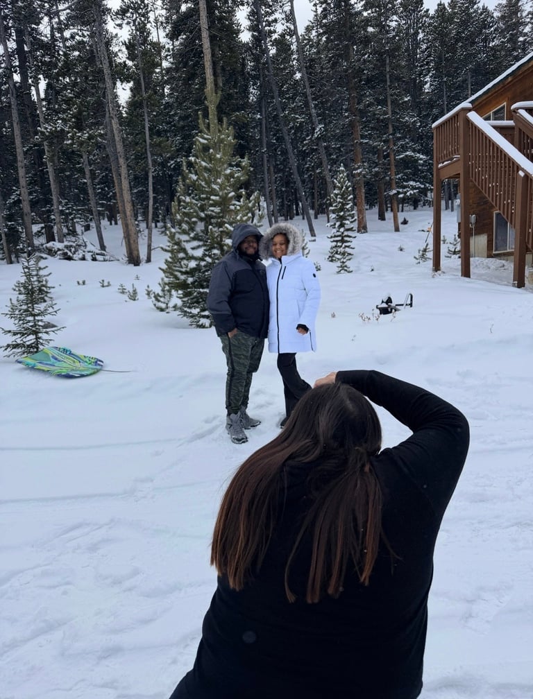 Person with long dark hair photographing two people standing in snowy forest landscape with cabin and evergreen trees