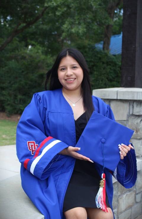 Young woman in blue graduation gown holding cap, smiling at camera with green trees and concrete wall in background