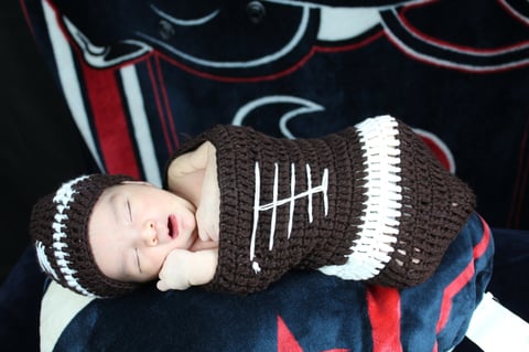 Newborn baby sleeping while wearing a brown knit football costume with white and red stripes