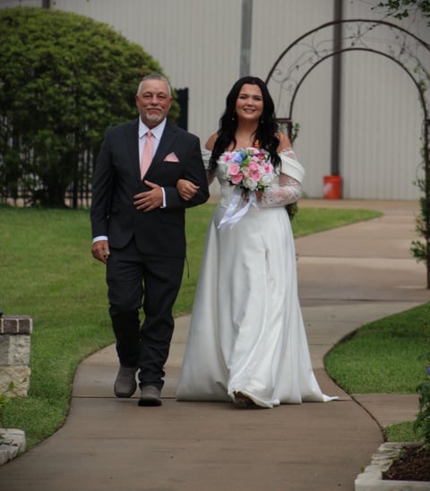 Bride in white wedding dress holding pink bouquet walking down garden pathway