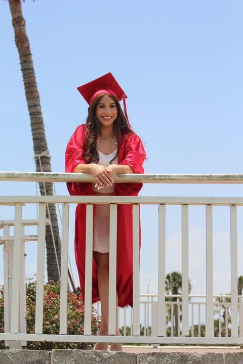 Woman in red graduation gown and cap leaning on a white railing overlooking palm trees and blue sky