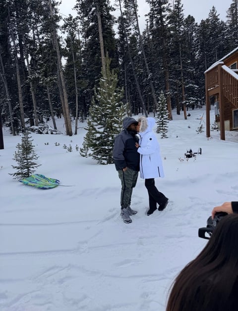 Two people walking in deep snow with a snowy forest and wooden cabin in the background