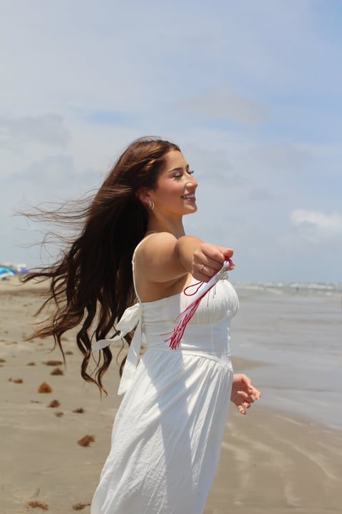 Woman in white dress with long dark hair holding a pink tassel on a sandy beach with ocean in background