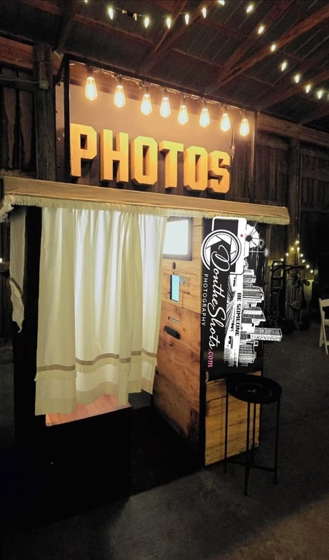 Vintage-style photo booth with yellow PHOTOS sign, white curtain entrance, and warm string lights in an indoor venue