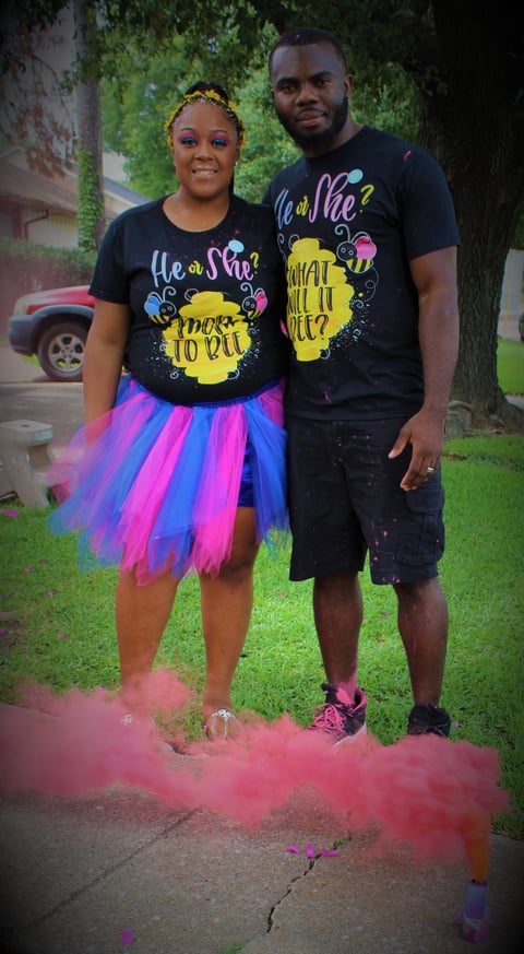 A couple wearing matching black graphic tees poses outdoors surrounded by pink powder, woman in colorful tutu skirt with gold headpiece