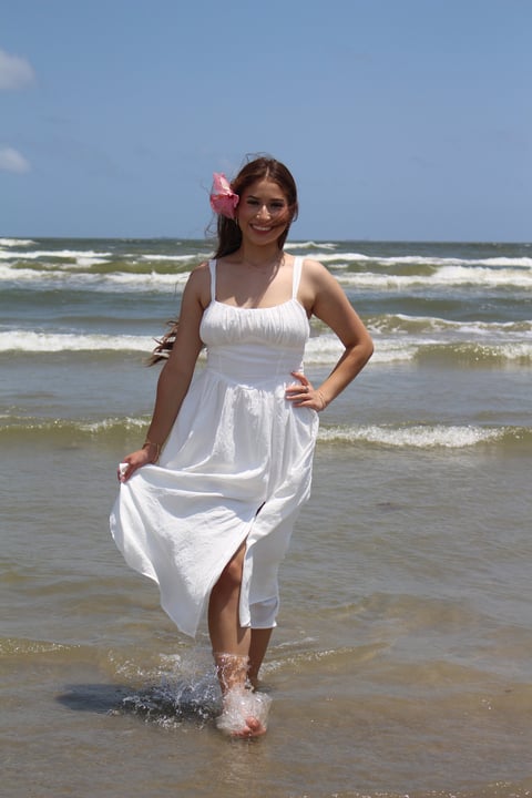 Woman in white sundress and sunglasses with pink flower in hair standing in the ocean waves on a beach