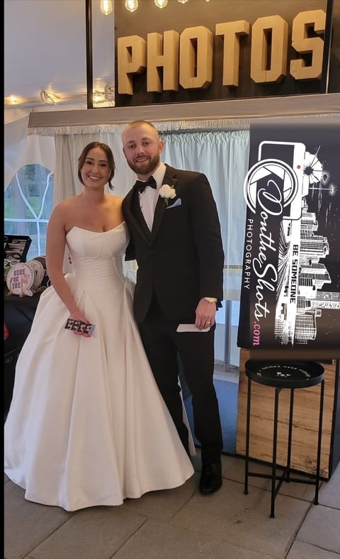 Bride in white strapless gown and groom in black tuxedo pose together at a photo booth at a wedding reception
