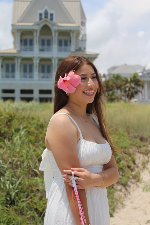Woman in white dress with pink flower in hair smiling outdoors with historic mansion in background