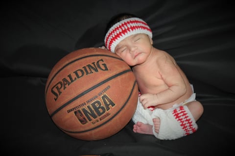 Newborn baby wearing red and white striped hat and diaper leaning against NBA basketball