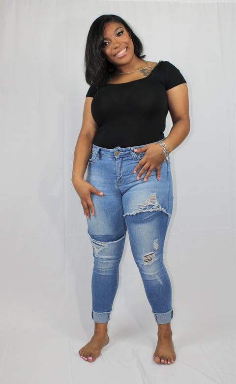 Woman wearing black short-sleeve top and distressed blue jeans, posing barefoot against white background