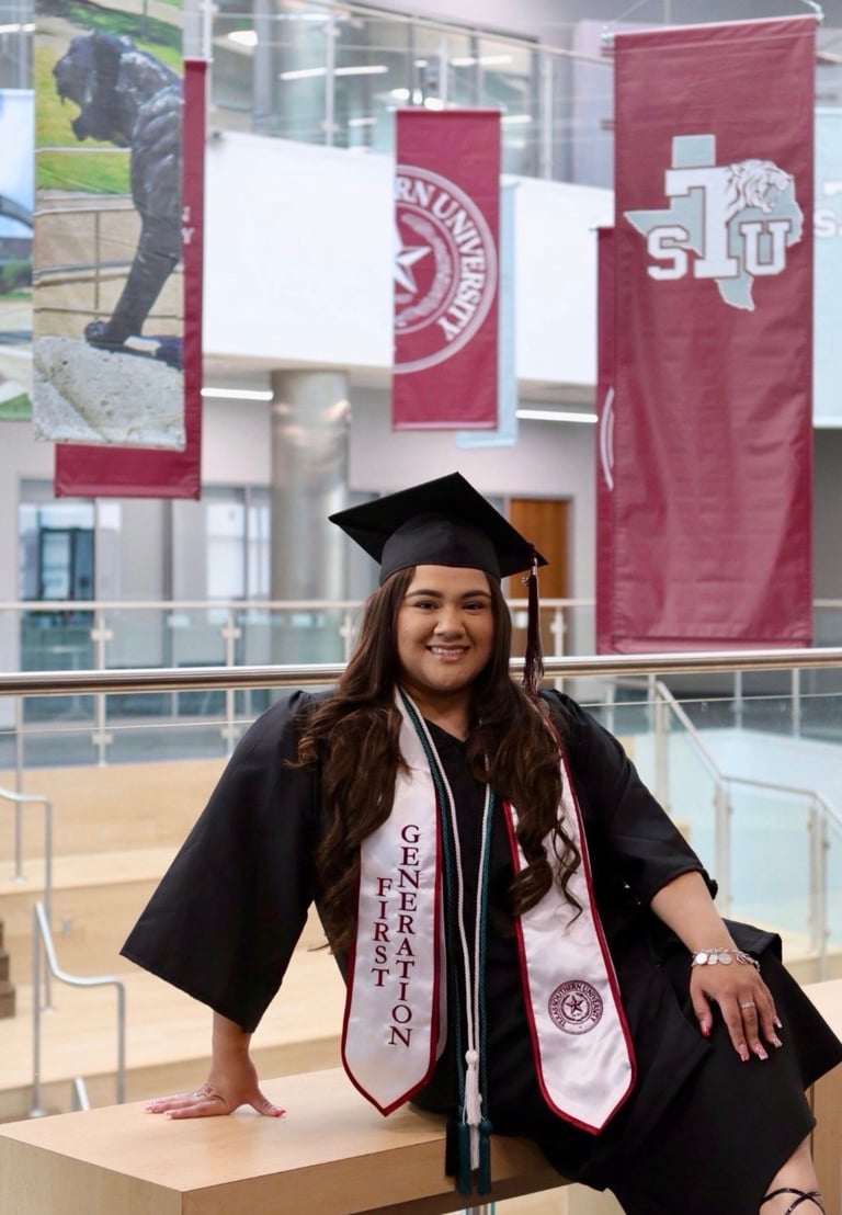 A graduate in black cap and gown with red sash stands in front of university banners at an indoor campus location
