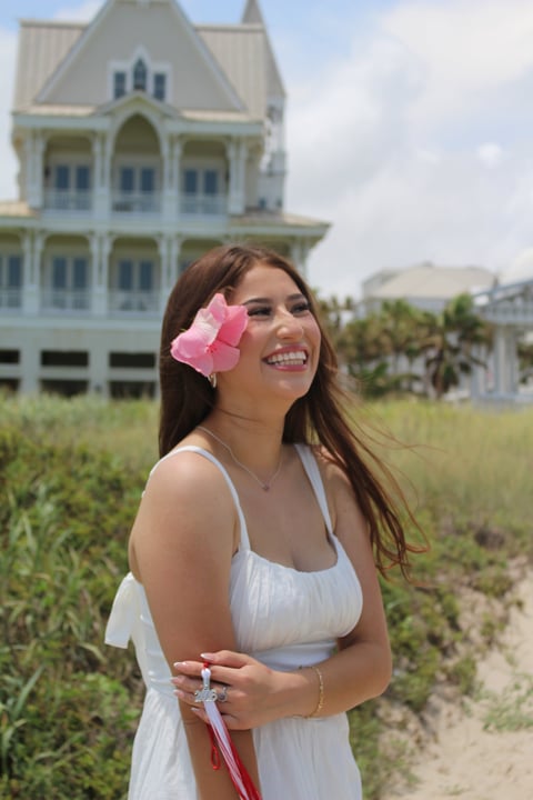 Smiling woman in white dress with pink hair bow posing in front of Victorian mansion