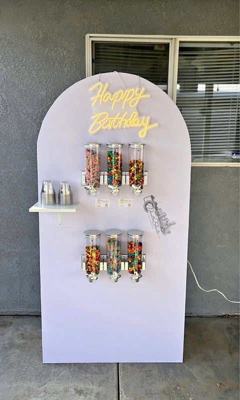 White arch-shaped candy dispenser display with yellow Happy Birthday neon sign and clear dispensers filled with colorful candies