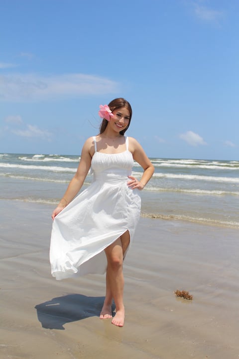Woman in white sundress with pink flower hair clip standing on beach with ocean waves in background