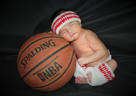 Newborn baby wearing red and white striped hat and diaper leaning on NBA basketball against dark background