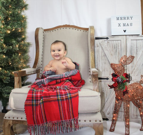 Baby sitting in upholstered chair wrapped in red plaid blanket with Christmas decorations and Merry Xmas sign in background