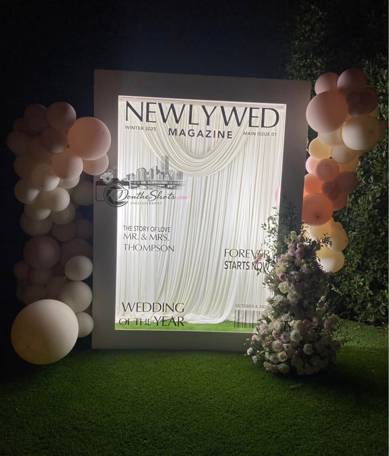 Large magazine-style wedding display frame surrounded by neutral and metallic balloons and floral arrangements on green artificial grass