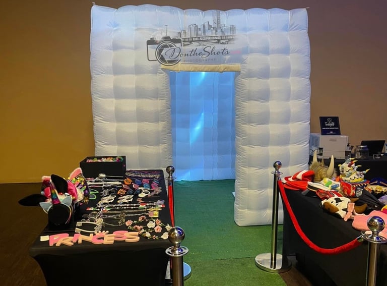 Inflatable photo booth with branded backdrop displaying wedding photography logo, flanked by product display tables with floral arrangements and equipment