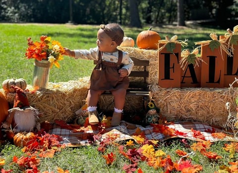 Toddler in brown outfit sits on hay bales surrounded by fall decorations, pumpkins, and colorful leaves in a garden setting.