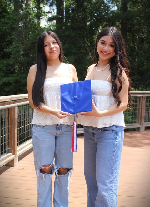Two young women in white tank tops holding a blue graduation cap on a wooden deck with green trees in the background