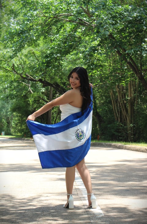 Woman holding El Salvador flag on tree-lined road, wearing white top and ankle boots