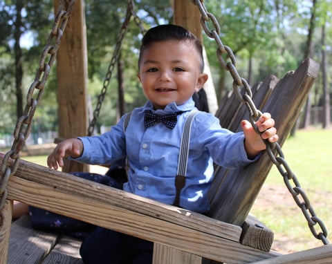 Toddler boy in blue shirt and bow tie smiling on a wooden swing in a park