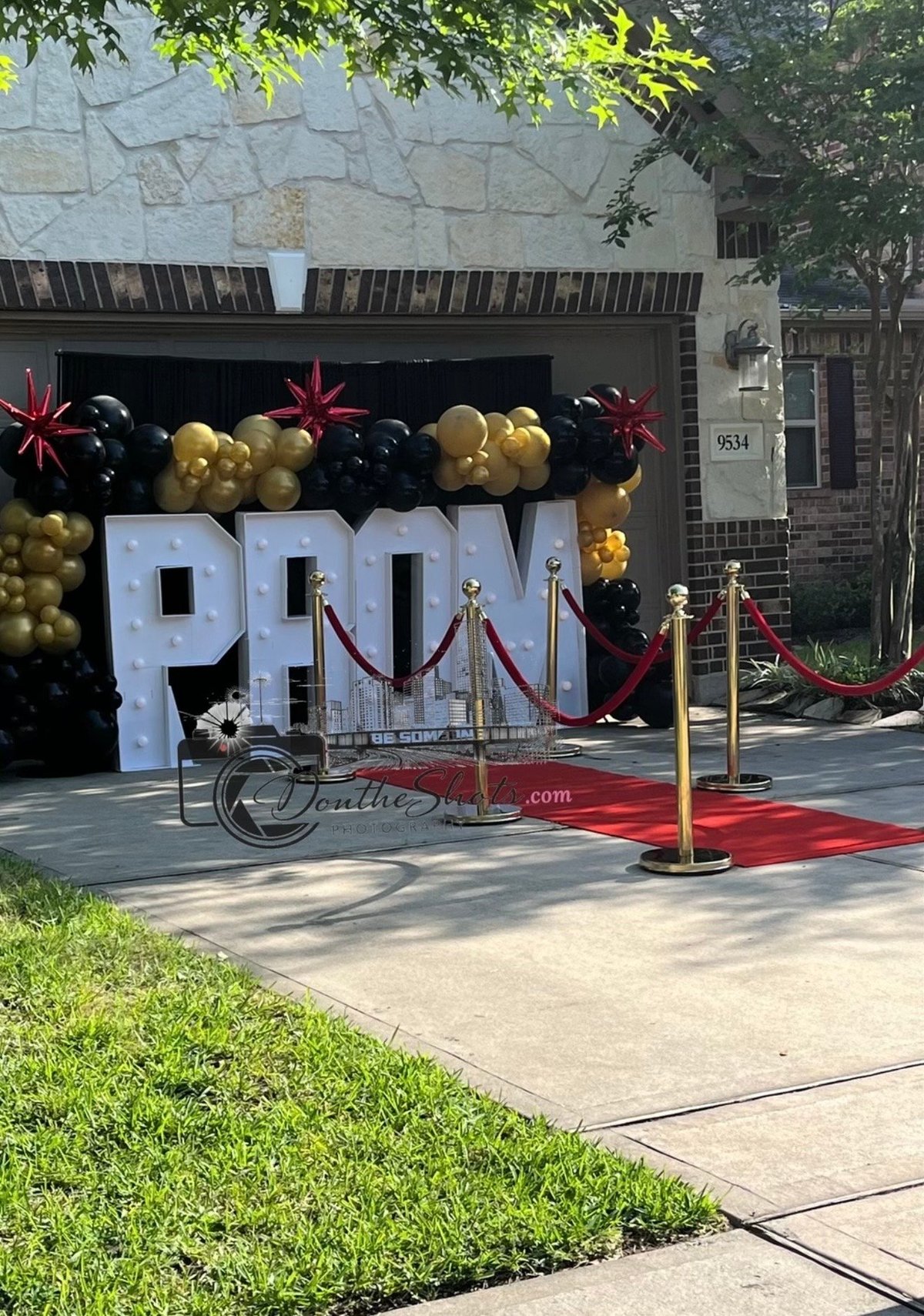 Storefront decorated with large white PROM letters, gold and black balloons, red carpet, velvet ropes, and a vintage bicycle in front of a brick building.