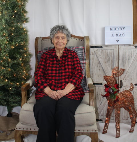 Elderly woman in red plaid shirt sitting in a chair with Christmas decorations, including a decorated tree and Merry Xmas sign behind her