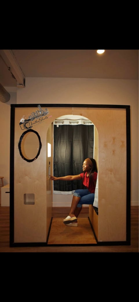 Woman in red shirt inside a wooden photo booth with black curtain backdrop and storefront opening