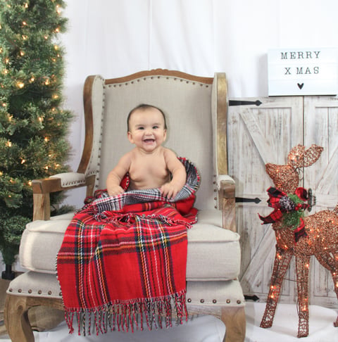 Smiling baby sits in upholstered chair wrapped in red plaid blanket with Christmas decorations and reindeer display in background